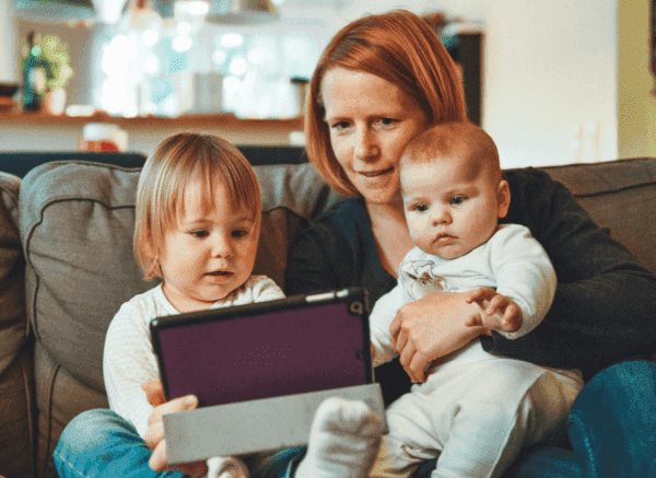 Woman sitting with a toddler and a baby looking at a tech device.